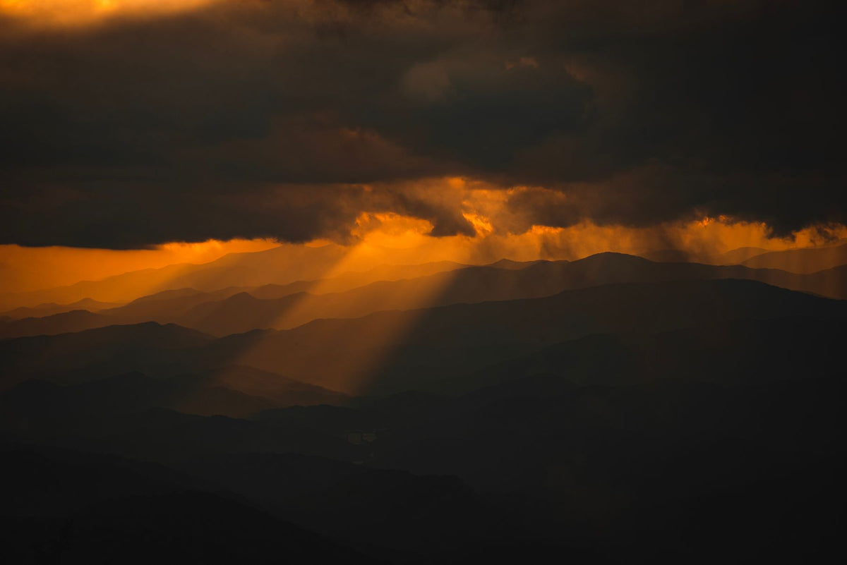 A single shaft of last light falling through cloud onto layered mountain ridges at dusk
